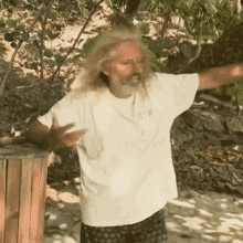 a man with long hair and a beard is standing on a beach with his arms outstretched and a white shirt on .