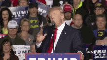 a man in a suit and tie is speaking into a microphone in front of a crowd with a sign that says trump