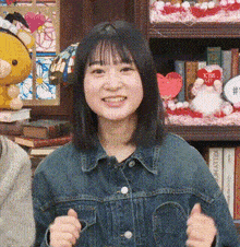 a woman in a denim jacket is giving a thumbs up in front of a bookshelf filled with stuffed animals .