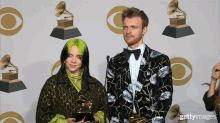 a woman with green hair and a man in a suit stand next to each other in front of a wall of grammy trophies