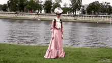 a woman in a pink dress and hat is standing on a lush green field next to a river .