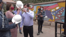 a man wearing a mask holds a white hard hat in front of a wall that says mediabancot on it