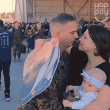 a man in a military uniform holds a baby while a woman kisses him in front of a sign that says hop pillu