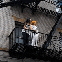 two women are standing on a fire escape on a brick building .