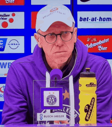 a man in a purple sweatshirt stands in front of a microphone next to a yellow water bottle that says neeraton