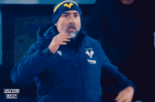 a man wearing a blue jacket and a yellow hat with the letter v on it shouts