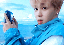 a young man in a blue jacket is holding a cell phone in front of the ocean