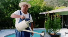 a man in overalls and a hat is watering plants with a watering can in front of a pool .