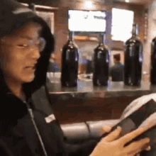 a man in a hooded jacket is reading a book in front of bottles of wine on a counter .