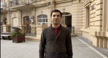 a man in a grey sweater stands in front of a building with a sign on it that says ' a '