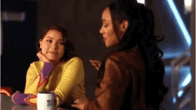 two women are sitting at a table with a cup of coffee and talking .