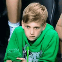 a young boy wearing a green hoodie is sitting in a stadium holding a green towel and a pen .