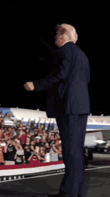 a man in a suit stands on a stage in front of a crowd with a plane in the background