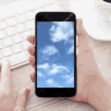 a person is typing on a keyboard while holding a cell phone with a picture of clouds on the screen .