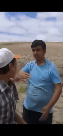 two men are standing next to each other in a field . one of the men is wearing a blue shirt and a white hat .