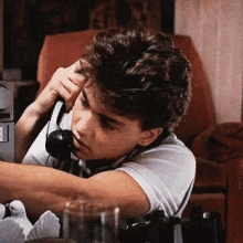 a young man is talking on a telephone while sitting at a table with a glass in front of him