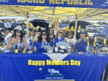 a group of people are sitting at a table under a tent with a blue table cloth that says happy mothers day .