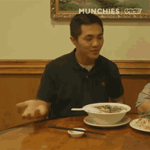 a man is sitting at a table with a bowl of soup and chopsticks and the words munchies visible in the corner