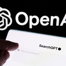 This image shows a close-up of a smartphone screen displaying the text “SearchGPT”; In the background, the OpenAI logo is prominently displayed,