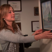 A woman (Jenna Fischer as Pam Beesly) removing a framed watercolor painting of an office building from the wall in a scene of "The Office."