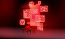 A person sits at a computer desk with numerous red screens around them. 