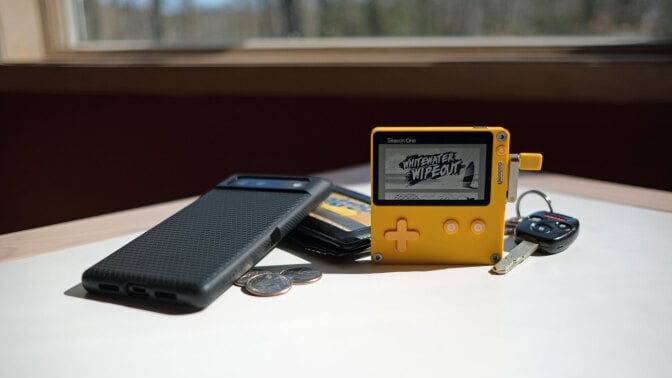 Playdate, a palm-sized gaming device, sits on a table surrounded by items typically carried in a pocket: A phone, a wallet, keys, and coins.