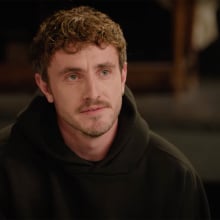 A man in a hoodie sits at a table, looking serious.