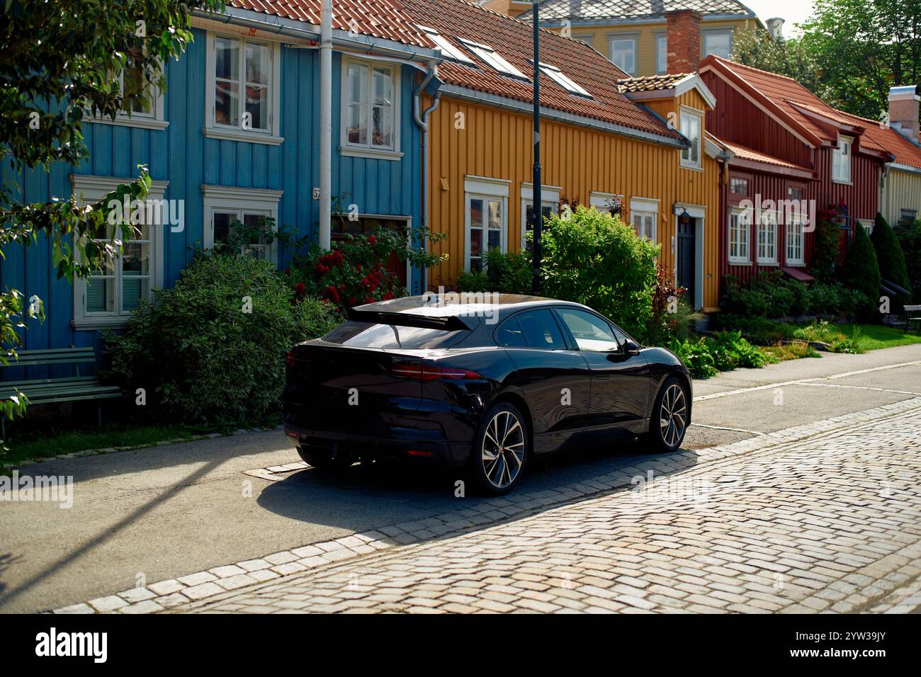 A sleek black electric car parked on a quaint street with colorful wooden houses on a sunny day, Trondheim, Norway Stock Photo