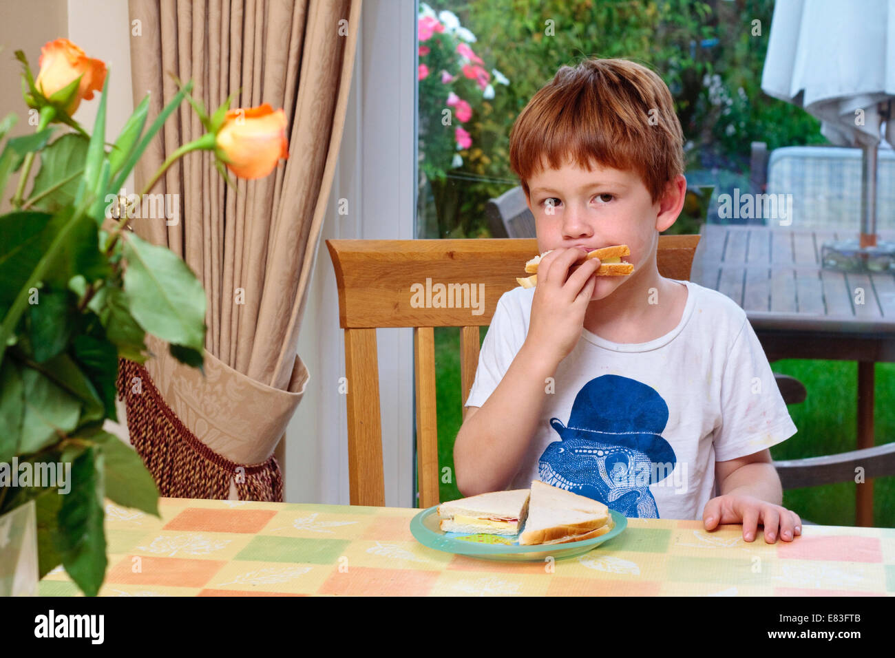 A young boy sat at a table eating a sandwich Stock Photo