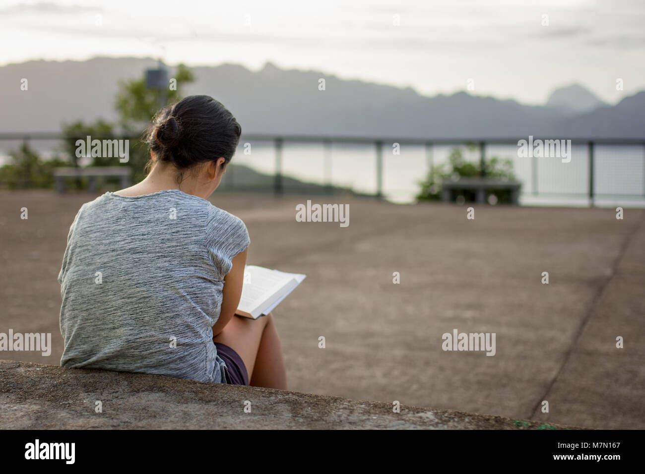 A young woman concentrates reading here hard back book whilst sat on some concrete steps. Stock Photo