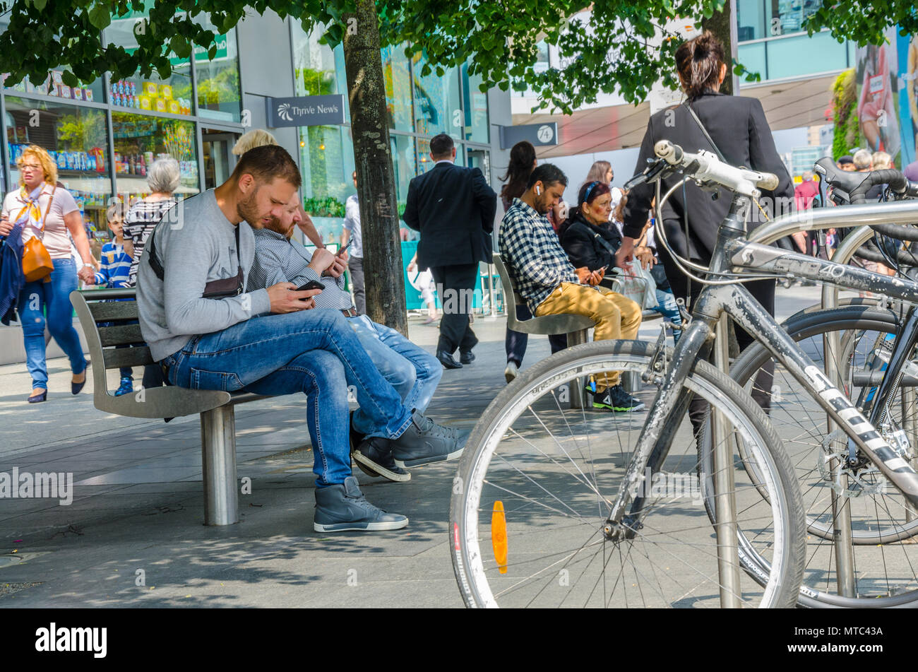 Bicycles parked in a bicycle rack next to benches with people sat preoccupied with their mobile phones, Stock Photo