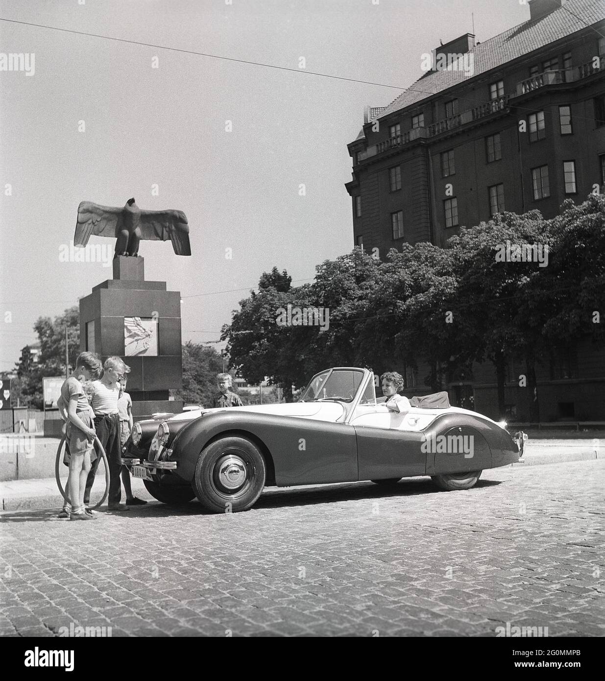 Fahren in den 1950er Jahren. Eine junge Frau in einem offenen Cabriolet des Jaguar XK120. Ein britischer Sportwagen, der als futuristisch anmutet. Die Zahlen 120 geben die Höchstgeschwindigkeit des Fahrzeugs an, 120 mph/193 km/h. Das Auto war ein Zweisitzer mit einer Aluminium-Chasse. Es wurde in insgesamt 12078 Autos hergestellt. Auf der Straße vom Flugdenkmal von Carl Milles auf Karlaplan in Stockholm abgebildet. Eine Hommage an schwedische Piloten und geflügelte Pioniere. Schweden 1953. ref. BL108-12 Stockfoto