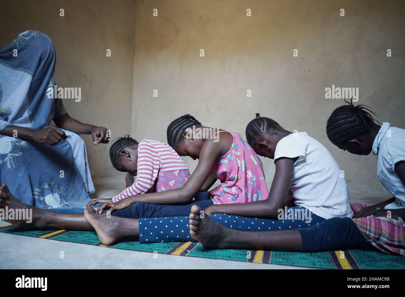 Eine Gruppe junger afrikanischer Kinder sitzt auf dem Boden und bildet eine Linie vor dem Mann mit Kopfbedeckung. Rituale; Traditionen; Konzept der kulturellen Vielfalt Stockfoto