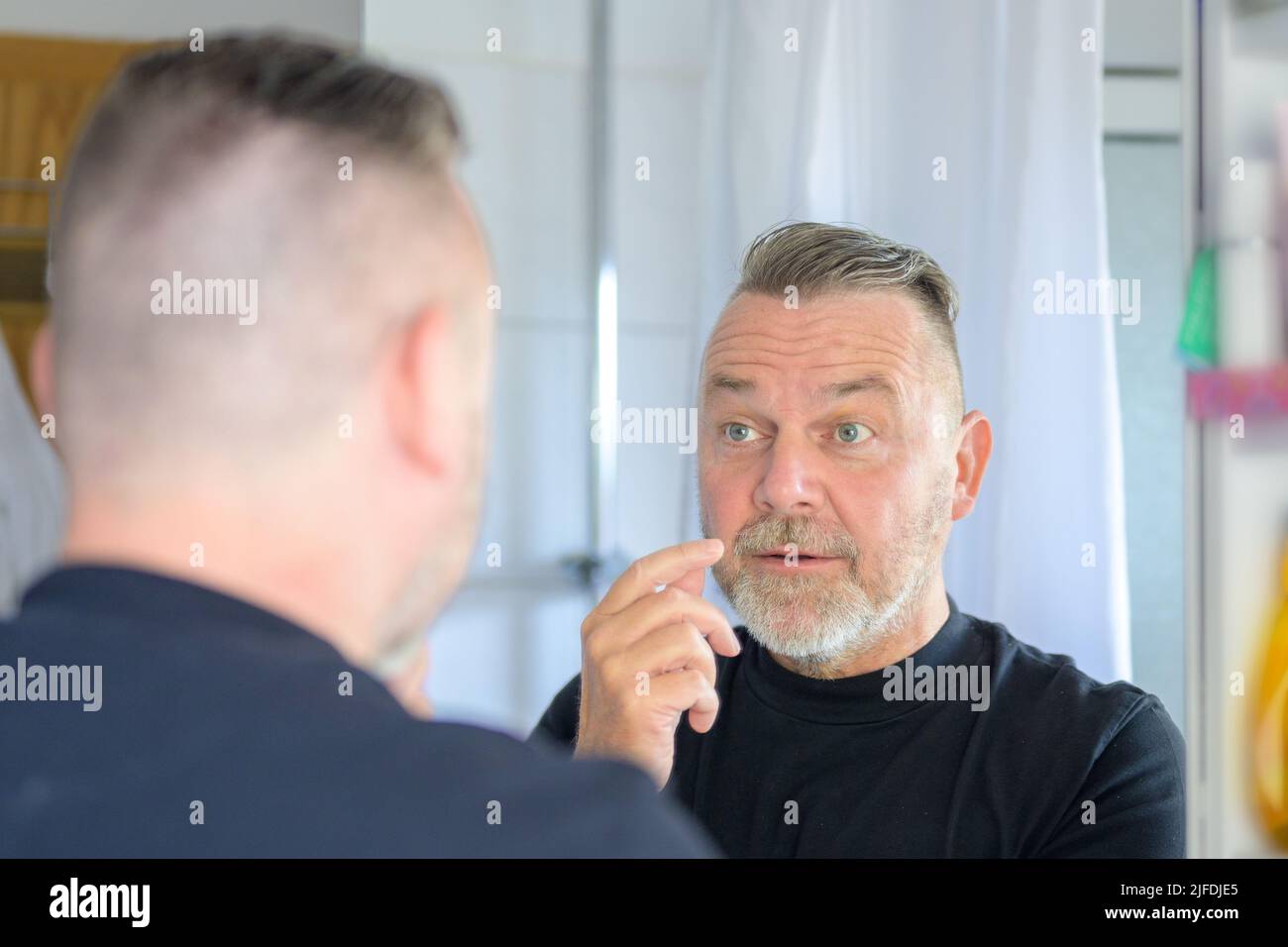 Bestürzter Mann mittleren Alters, der die fleischigen Taschen unter seinen Augen ansah, mit Blick auf seine Spiegelung im Spiegel Stockfoto