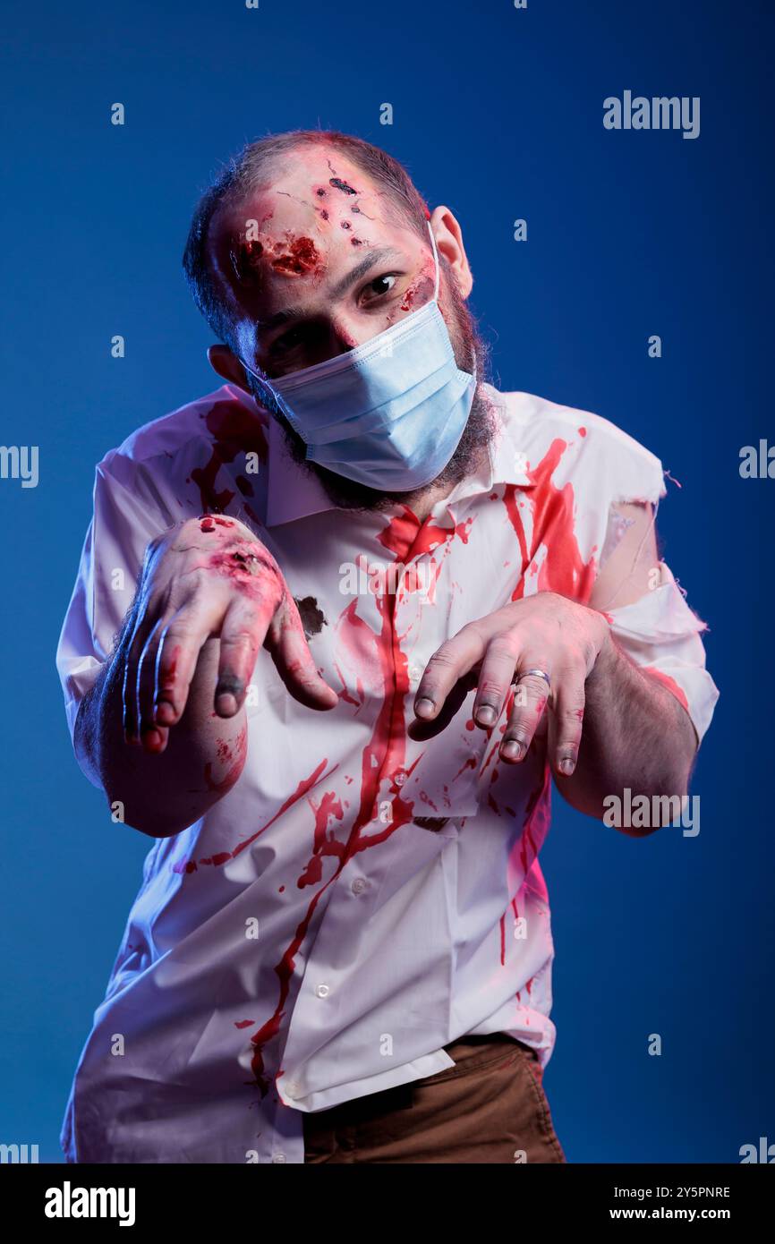 Porträt eines Mannes, der als finstere Zombie verkleidet ist, bedeckt mit Blut, der Gesichtsmaske für halloween-Event trägt. Person trägt Gore Make-up und gibt vor, untote Kreatur während des Virusausbruchs zu sein, Studiohintergrund Stockfoto