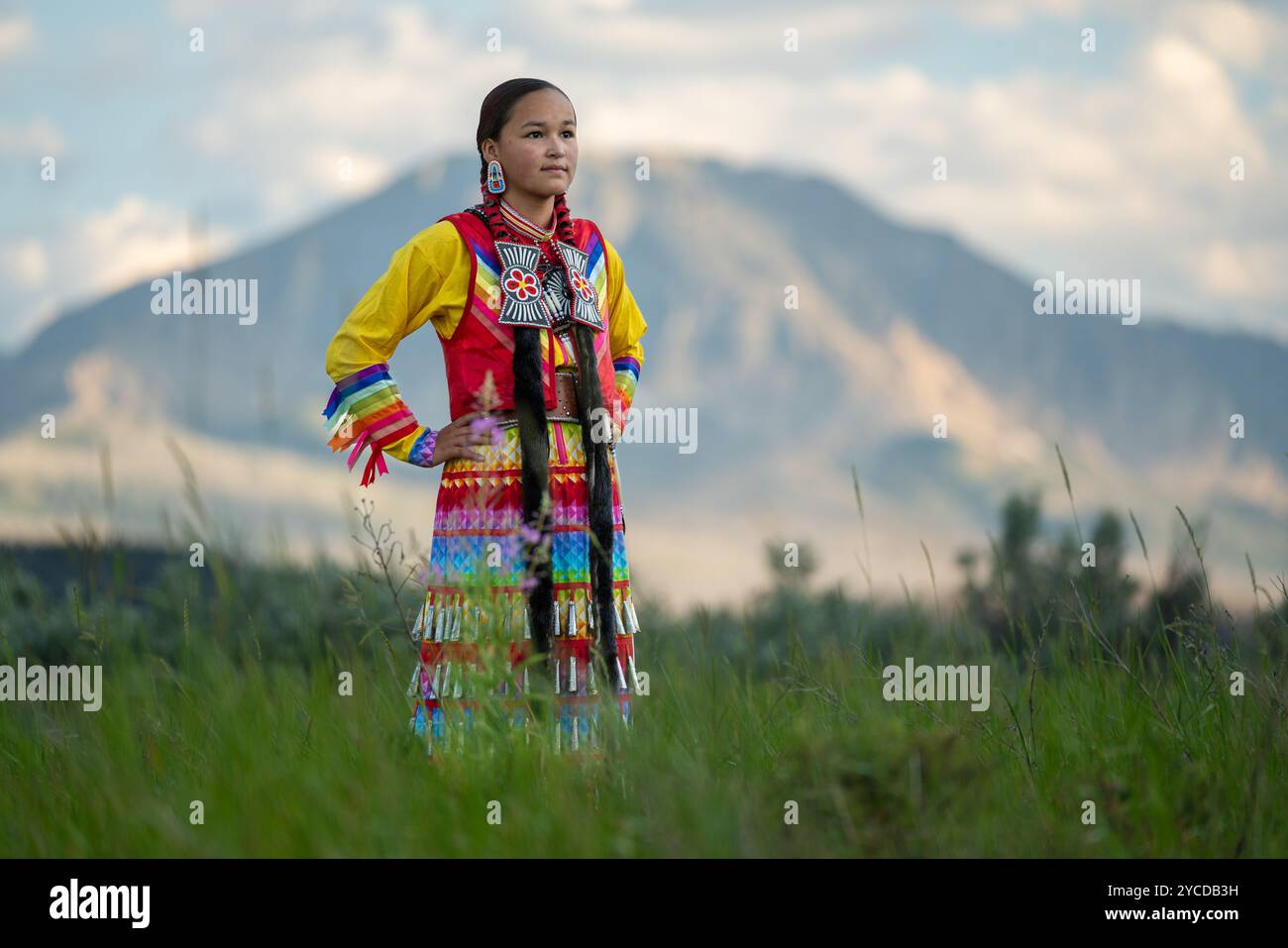 Ein junges Mädchen in traditioneller Ureinwohnerkleidung steht stolz auf einem grasbewachsenen Feld mit Bergen im Hintergrund und feiert das kulturelle Erbe und Stockfoto