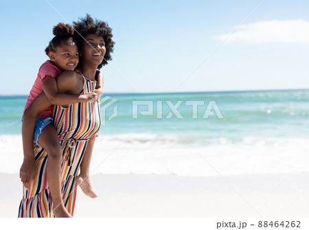 Smiling african american mother giving piggyback ride to daughter at beach on sunny day 88464262